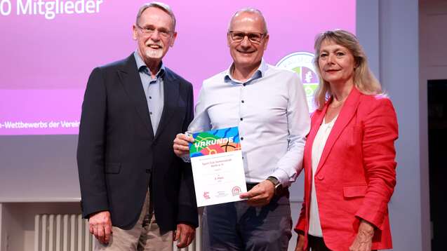 Drei Erwachsene stehen zusammen auf einer B&uuml;hne und halten eine Urkunde des Vereinswettbewerbs in der Hand.