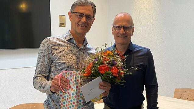 Zwei Männer stehen lächelnd nebeneinander, einer hält einen Blumenstrauß und die andere eine Geschenktüte mit Karte.