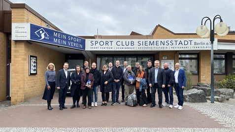 Gruppe von Personen vor dem Eingang des Sport Club Siemensstadt in Berlin, mit Schild und verschiedenen Pflanzen im Hintergrund.