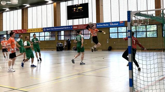 Aktion im Handballspiel: Spieler in orange springt zum Torwurf, während grüne Spieler und Torwart beobachten.