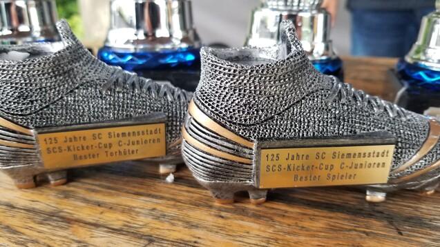 Zwei Fußballschuhe aus Metall mit Plaketten für „Bester Torhüter“ und „Bester Spieler“ auf einem Holztisch.