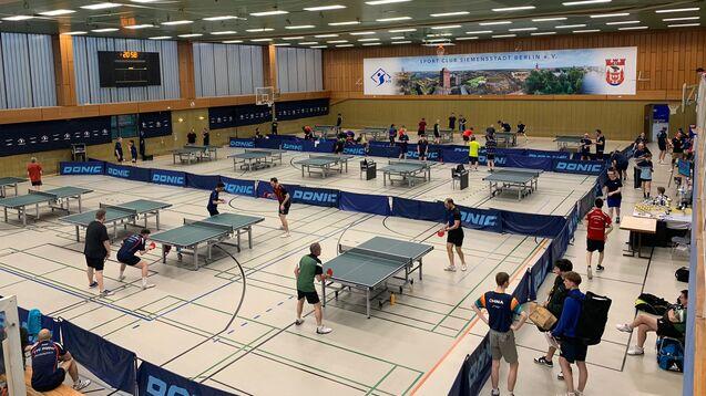 Aktive Tischtennisspieler in einer Halle mit mehreren Tischen, Banden und einem Zeitgeber im Hintergrund.