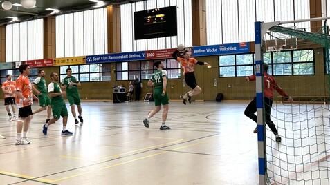 Aktion im Handballspiel: Spieler in orange springt zum Torwurf, während grüne Spieler und Torwart beobachten.