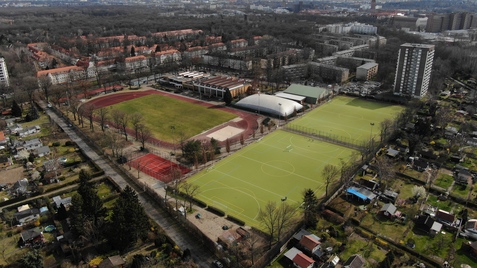 Luftaufnahme eines Sportgeländes mit Tennisplätzen, Fußballfeld und Wohngebäuden in der Umgebung.