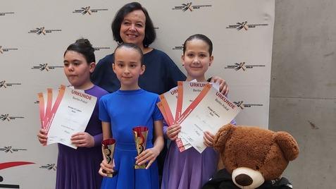 Drei Mädchen in Tanzkleidern halten Pokale und Urkunden, stehen mit einer Frau vor einem Banner des Tanzsportverbands.