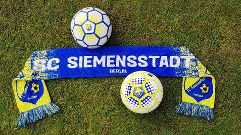 Zwei Fußbälle und ein blau-gelber Schal des ISC Siemensstadt liegen auf grünem Rasen.