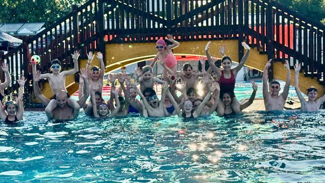 Gruppenshot von Kindern und Erwachsenen im Wasser, die fröhlich mit erhobenen Händen posieren, im Hintergrund eine Rutsche.