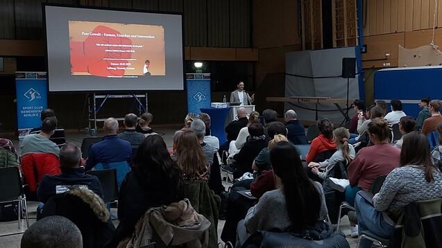 Vortrag zu Peer-Gewalt mit Projektorbildschirm und Publikum in einem Sportraum. Der Redner steht hinter einem Tisch.