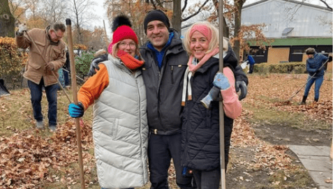 Drei Personen posieren auf einer herbstlichen Wiese, während im Hintergrund weitere Menschen Laub zusammenräumen.