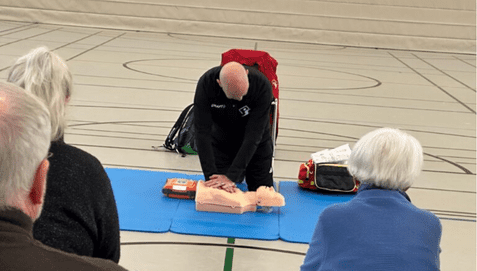Ein Trainer führt eine Herz-Lungen-Wiederbelebung an einer Übungs-Puppe vor, während Teilnehmer zuschauen.