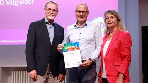 Drei Erwachsene stehen zusammen auf einer Bühne und halten eine Urkunde des Vereinswettbewerbs in der Hand.