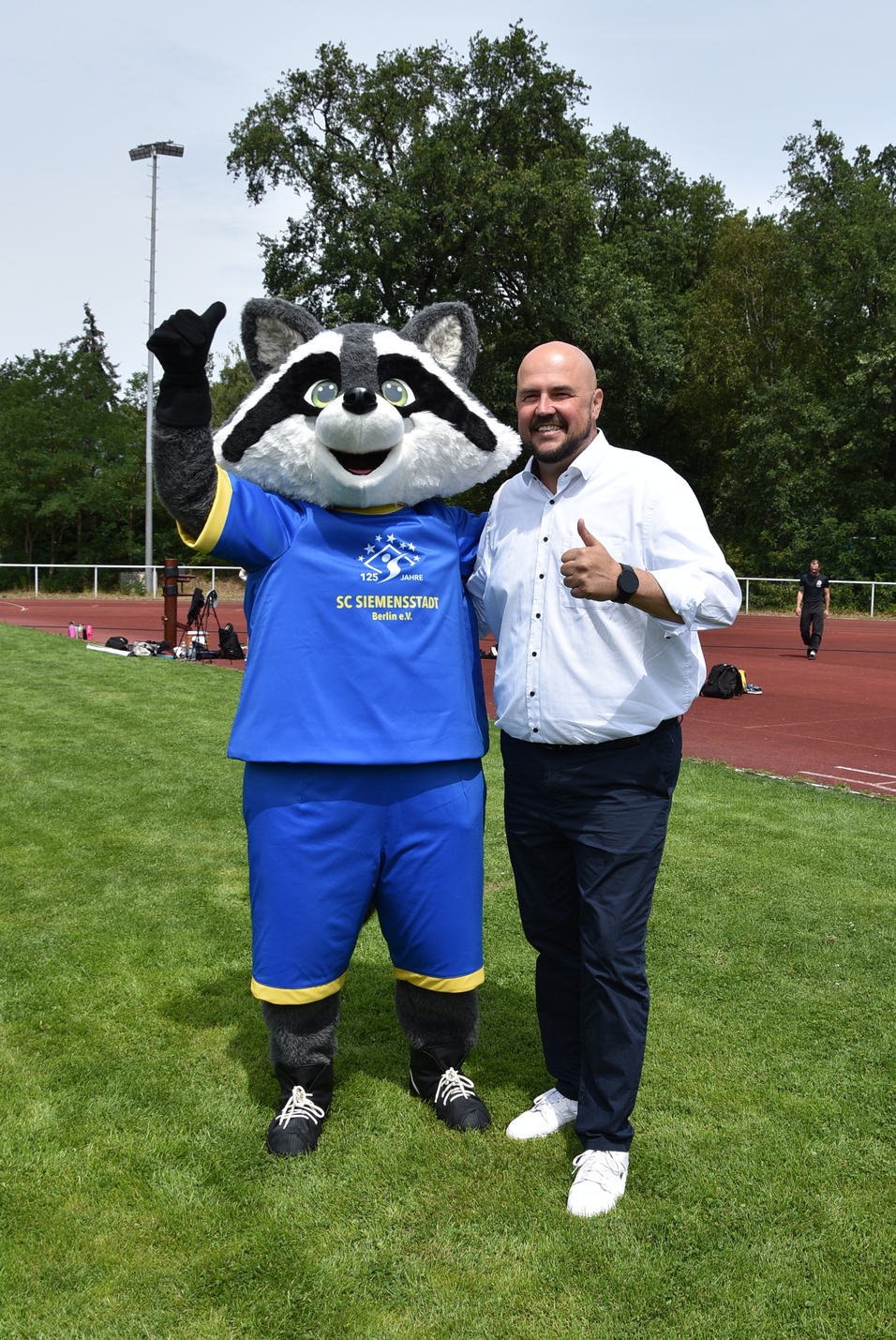 Mann in weißem Hemd und Maskottchen mit Waschbär-Kostüm posieren lächelnd auf einem Sportplatz.