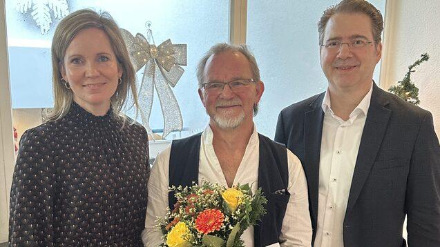 Drei Personen stehen in einem festlichen Raum, einer hält einen Blumenstrauß und eine Karte. Weihnachtsdekoration im Hintergrund.