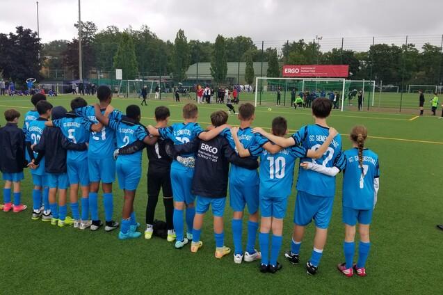 Gruppe von Kindern in blauen Fu&szlig;balltrikots, die Arm in Arm am Spielfeldrand stehen und ein Spiel beobachten.