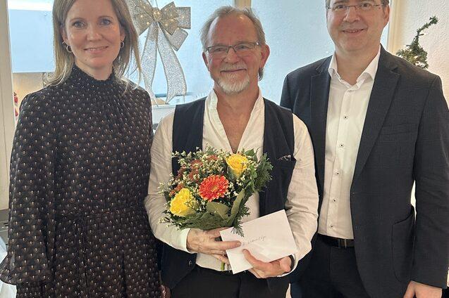 Drei Personen stehen in einem festlichen Raum, einer h&auml;lt einen Blumenstrau&szlig; und eine Karte. Weihnachtsdekoration im Hintergrund.