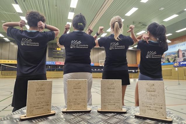 Vier Personen in T-Shirts mit Vereinslogo zeigen ihre Medaillen in einer Sporthalle. Auf einem Tisch stehen drei Pokale.