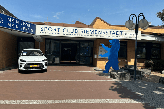 Eingangsbereich des Sport Club Siemensstadt in Berlin mit einem weißen Auto und einer blauen Bärenfigur.