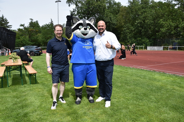 Drei Personen, darunter ein Maskottchen, posieren auf einem Sportplatz mit gr&uuml;ner Wiese im Hintergrund.
