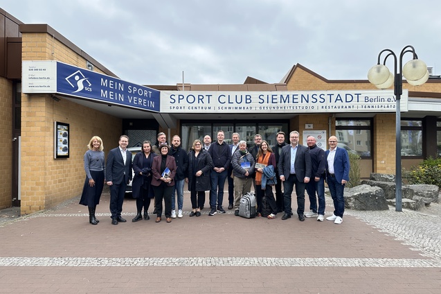 Gruppe von Personen vor dem Eingang des Sport Club Siemensstadt in Berlin, mit Schild und verschiedenen Pflanzen im Hintergrund.