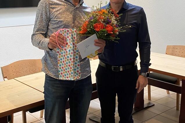 Zwei M&auml;nner stehen l&auml;chelnd nebeneinander, einer h&auml;lt einen Blumenstrau&szlig; und die andere eine Geschenkt&uuml;te mit Karte.