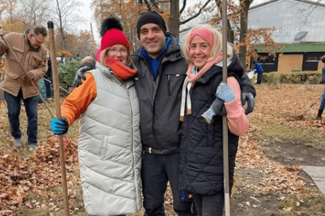 Drei Personen posieren auf einer herbstlichen Wiese, w&auml;hrend im Hintergrund weitere Menschen Laub zusammenr&auml;umen.