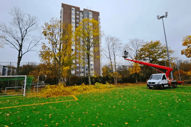 Rote Arbeitsb&uuml;hne mit einer Person hebt sich &uuml;ber einen Sportplatz, um Laub von B&auml;umen zu entfernen, umgeben von herbstlichen B&auml;umen.