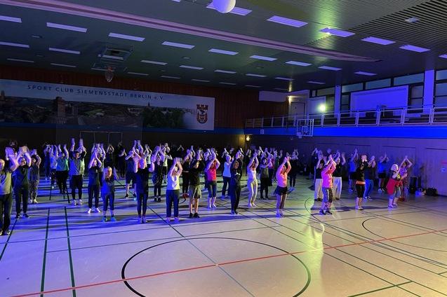 Gruppentanzveranstaltung in einer Sporthalle mit Teilnehmern, die synchron ihre Arme in die Luft heben.