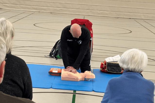 Ein Trainer f&uuml;hrt eine Herz-Lungen-Wiederbelebung an einer &Uuml;bungs-Puppe vor, w&auml;hrend Teilnehmer zuschauen.