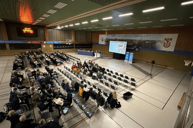 Versammlung in einer Sporthalle mit zahlreichen St&uuml;hlen und einer B&uuml;hne im Hintergrund. Zuschauer folgen der Pr&auml;sentation.