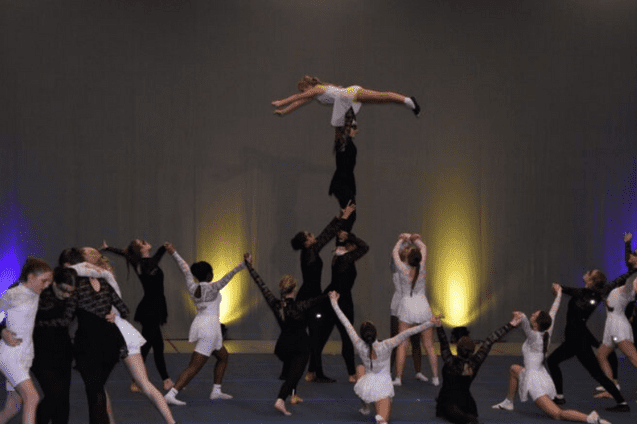 Tanzgruppe f&uuml;hrt akrobatische Elemente auf, mit einer T&auml;nzerin in der Luft &uuml;ber mehreren anderen T&auml;nzern.