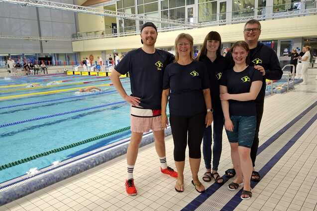 Gruppe von f&uuml;nf Schwimmern in Vereinskleidung steht am Beckenrand eines Schwimmbeckens w&auml;hrend eines Wettkampfs.