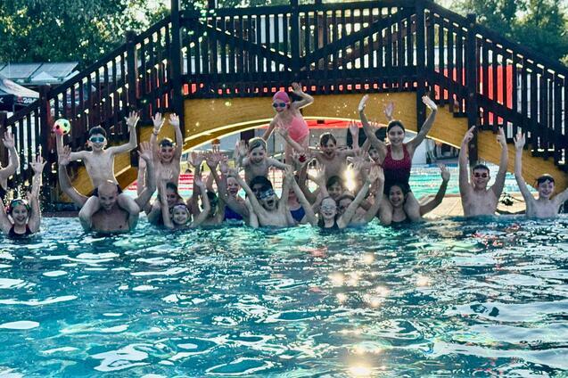 Gruppenshot von Kindern und Erwachsenen im Wasser, die fröhlich mit erhobenen Händen posieren, im Hintergrund eine Rutsche.