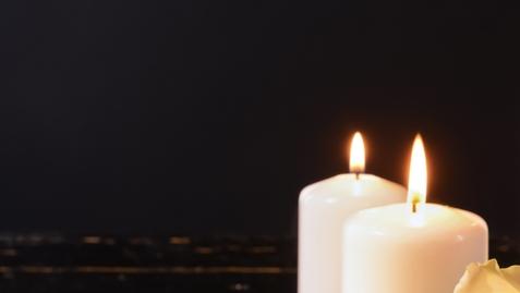 Zwei brennende, weiße Kerzen stehen vor einem dunklen Hintergrund.