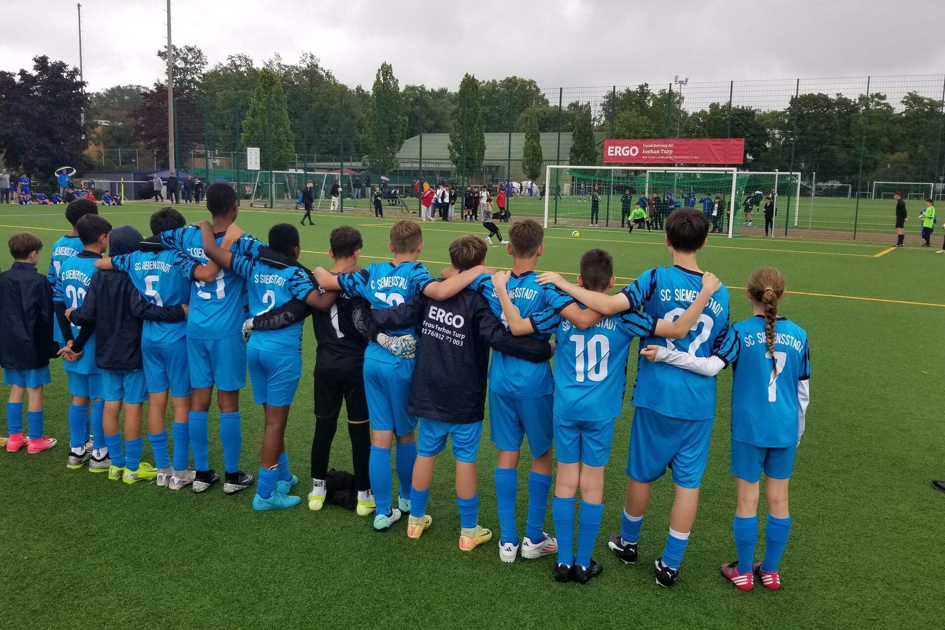 Gruppe von Kindern in blauen Fu&szlig;balltrikots, die Arm in Arm am Spielfeldrand stehen und ein Spiel beobachten.