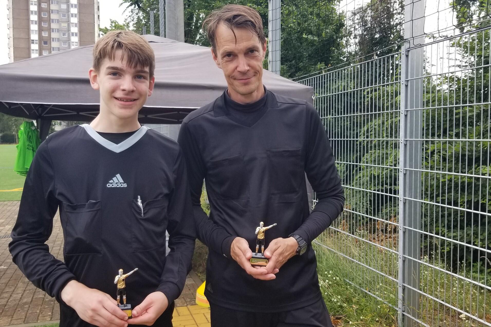 Zwei Fu&szlig;ballspieler in schwarzen Trikots halten Pokale in einer Au&szlig;enumgebung mit Sportplatz und Zelt im Hintergrund.