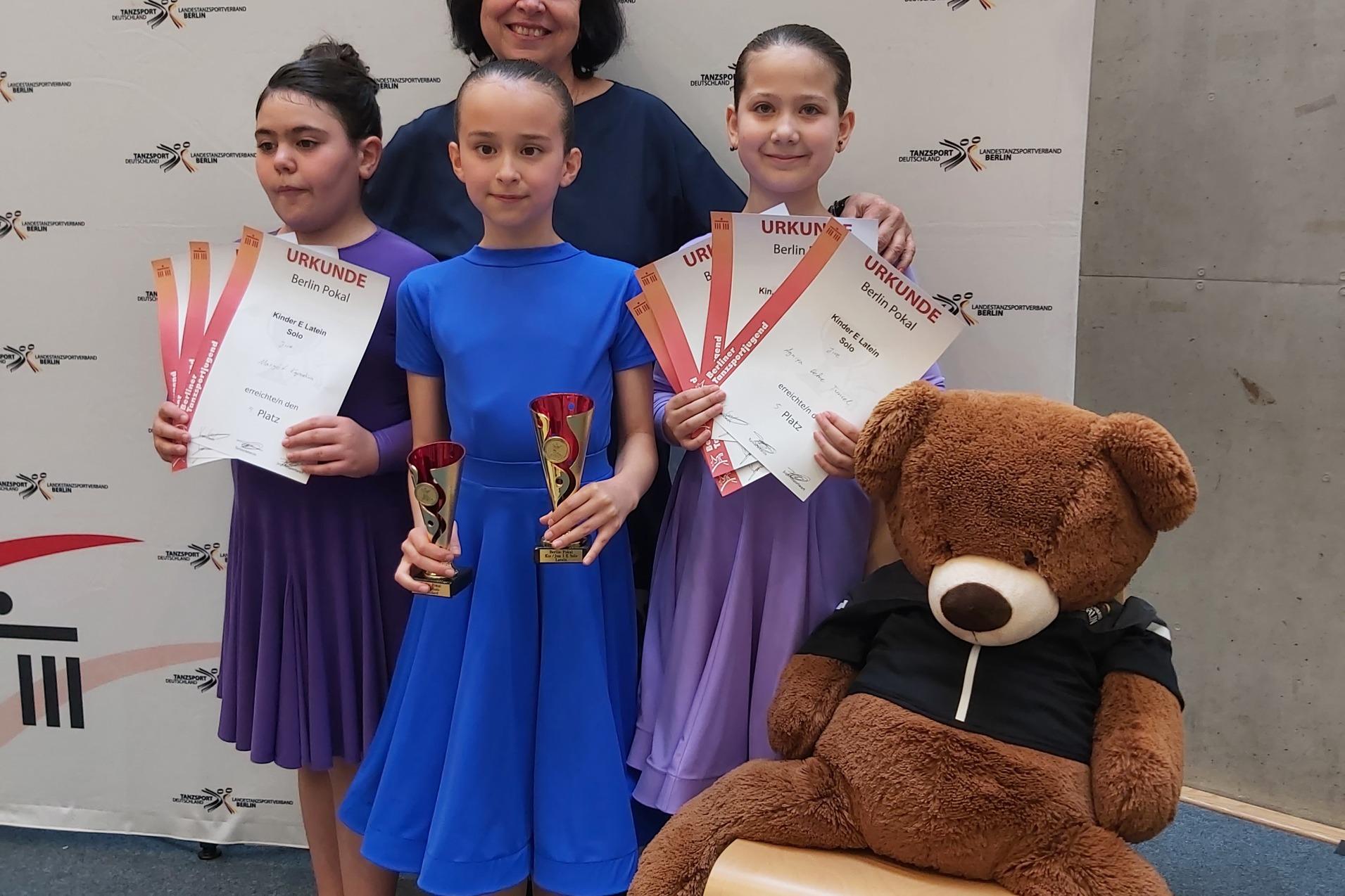 Drei M&auml;dchen in Tanzkleidern halten Pokale und Urkunden, stehen mit einer Frau vor einem Banner des Tanzsportverbands.