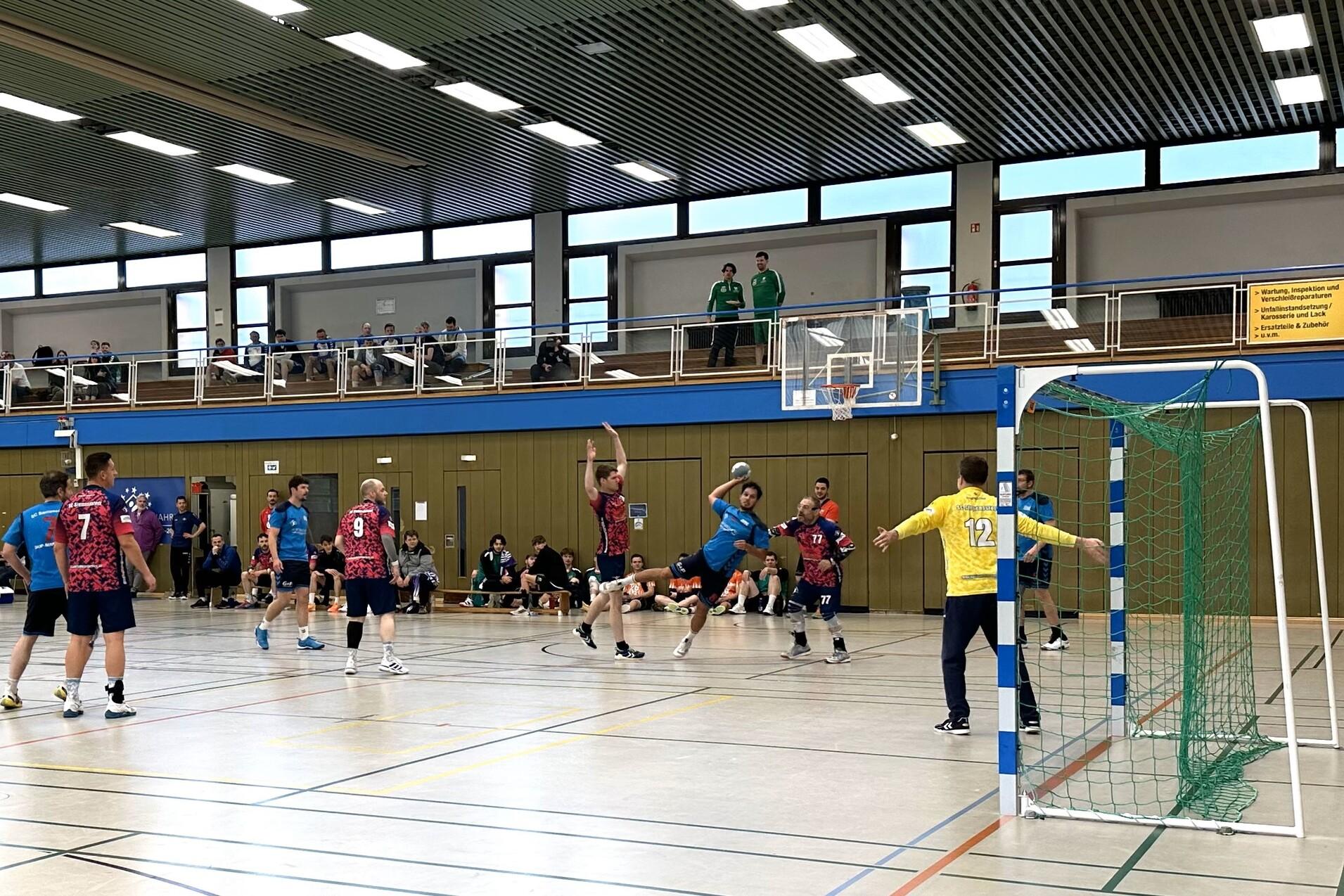 Handballsspieler in Aktion, ein Spieler springt zum Torwurf, Torwart bereit zur Abwehr. Zuschauer im Hintergrund.