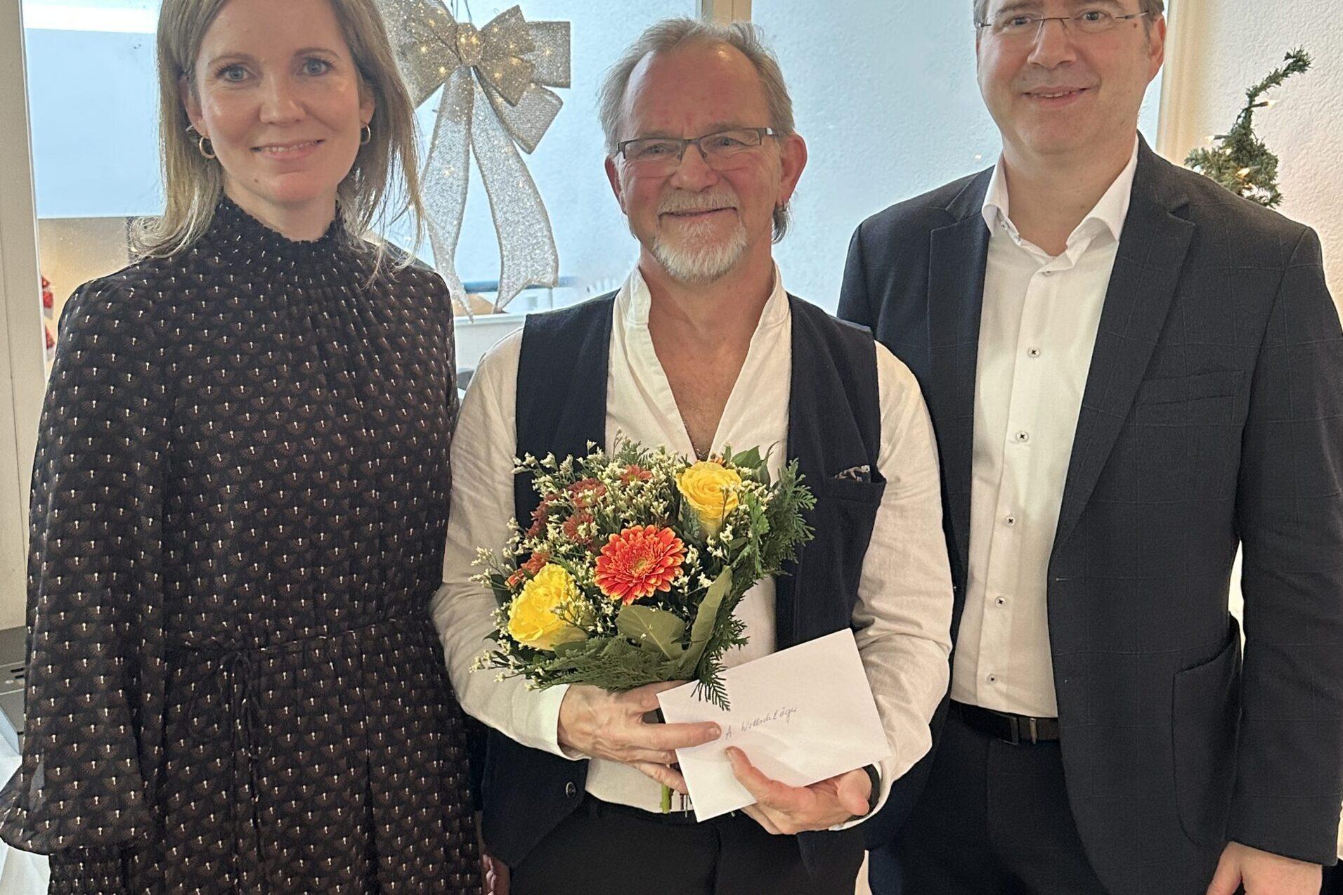 Drei Personen stehen in einem festlichen Raum, einer h&auml;lt einen Blumenstrau&szlig; und eine Karte. Weihnachtsdekoration im Hintergrund.