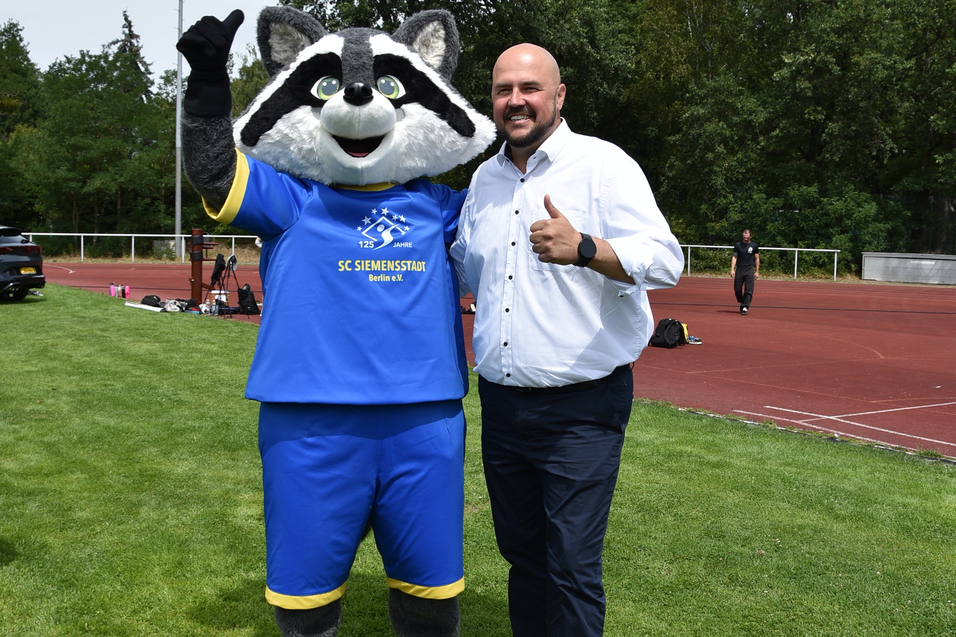 Mann in wei&szlig;em Hemd und Maskottchen mit Waschb&auml;r-Kost&uuml;m posieren l&auml;chelnd auf einem Sportplatz.