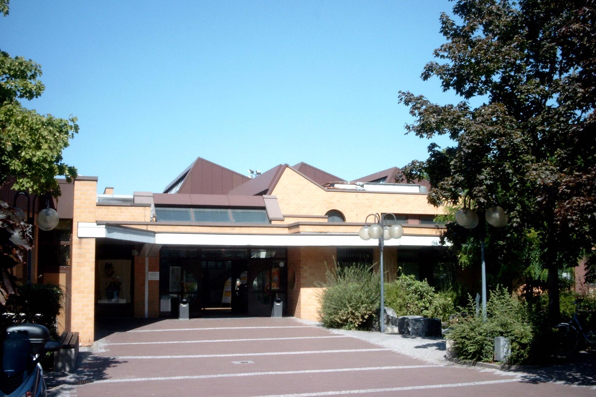 Eingangsbereich eines modernen Geb&auml;udes mit Glasfront, begr&uuml;ntem Umfeld und blauem Himmel.