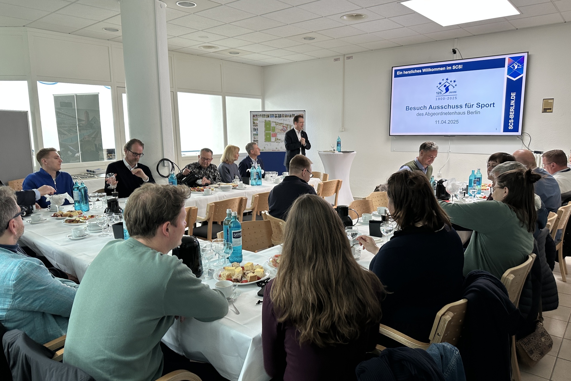 Gruppentreffen im SCSI mit Anwesenden am Tisch, Pr&auml;sentation im Hintergrund, Speisen und Getr&auml;nke auf dem Tisch.