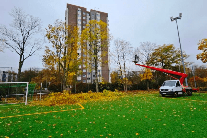 Rote Arbeitsb&uuml;hne mit einer Person hebt sich &uuml;ber einen Sportplatz, um Laub von B&auml;umen zu entfernen, umgeben von herbstlichen B&auml;umen.