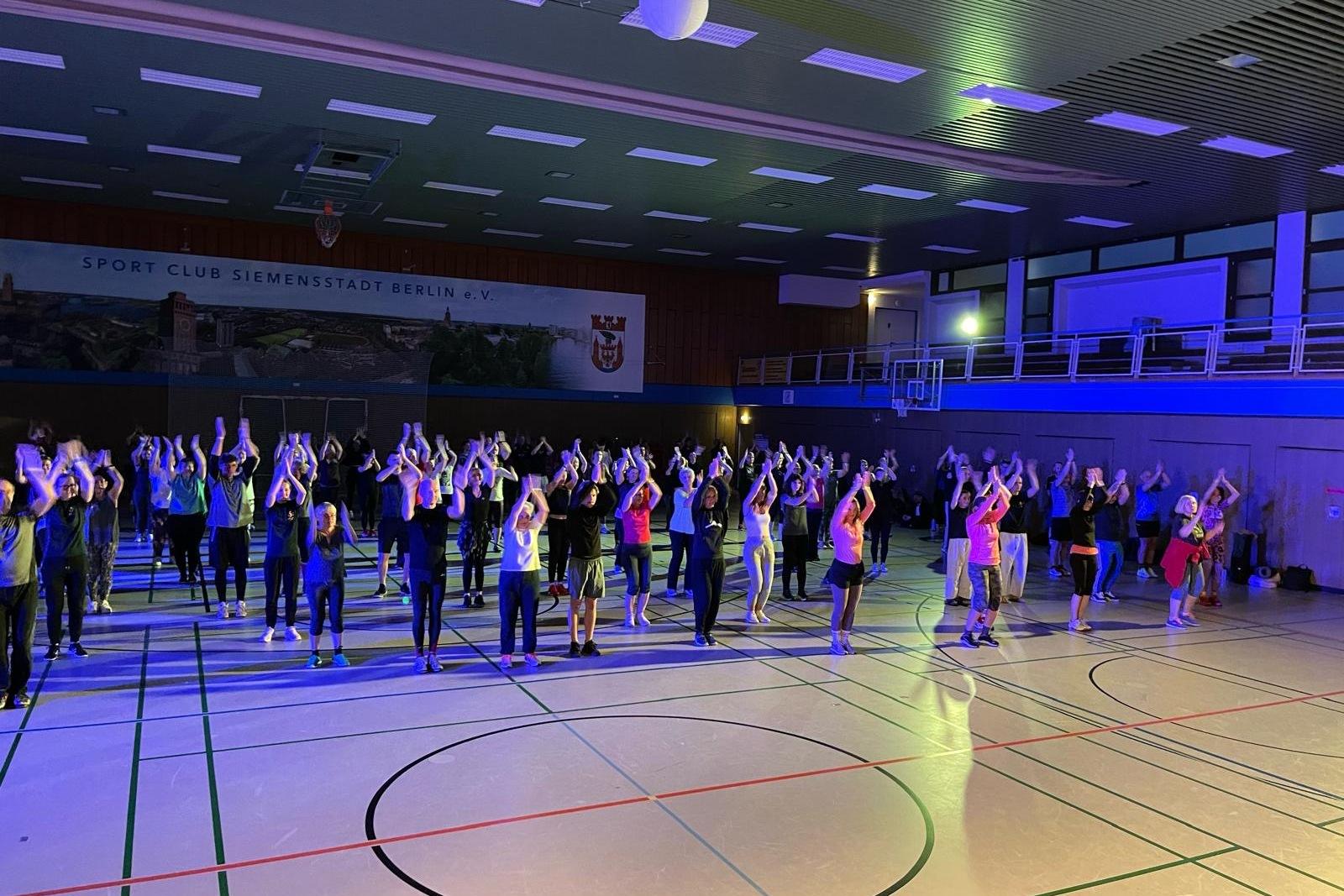 Gruppentanzveranstaltung in einer Sporthalle mit Teilnehmern, die synchron ihre Arme in die Luft heben.