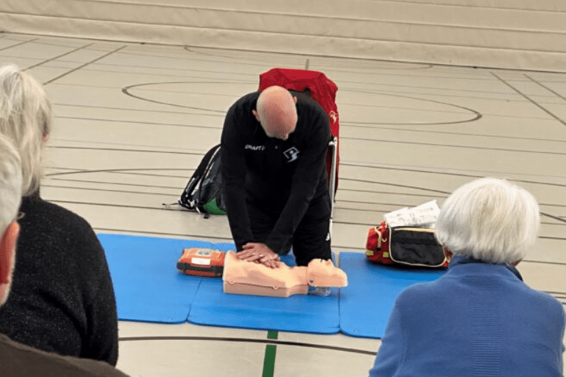 Ein Trainer f&uuml;hrt eine Herz-Lungen-Wiederbelebung an einer &Uuml;bungs-Puppe vor, w&auml;hrend Teilnehmer zuschauen.