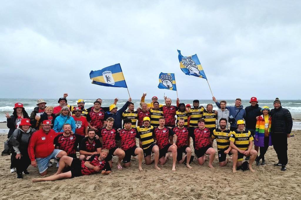 Gruppenantwortung von Rugbyspielern in Teamtrikots mit Fahnen am Strand, umgeben von Unterst&uuml;tzern in bunten H&uuml;ten.