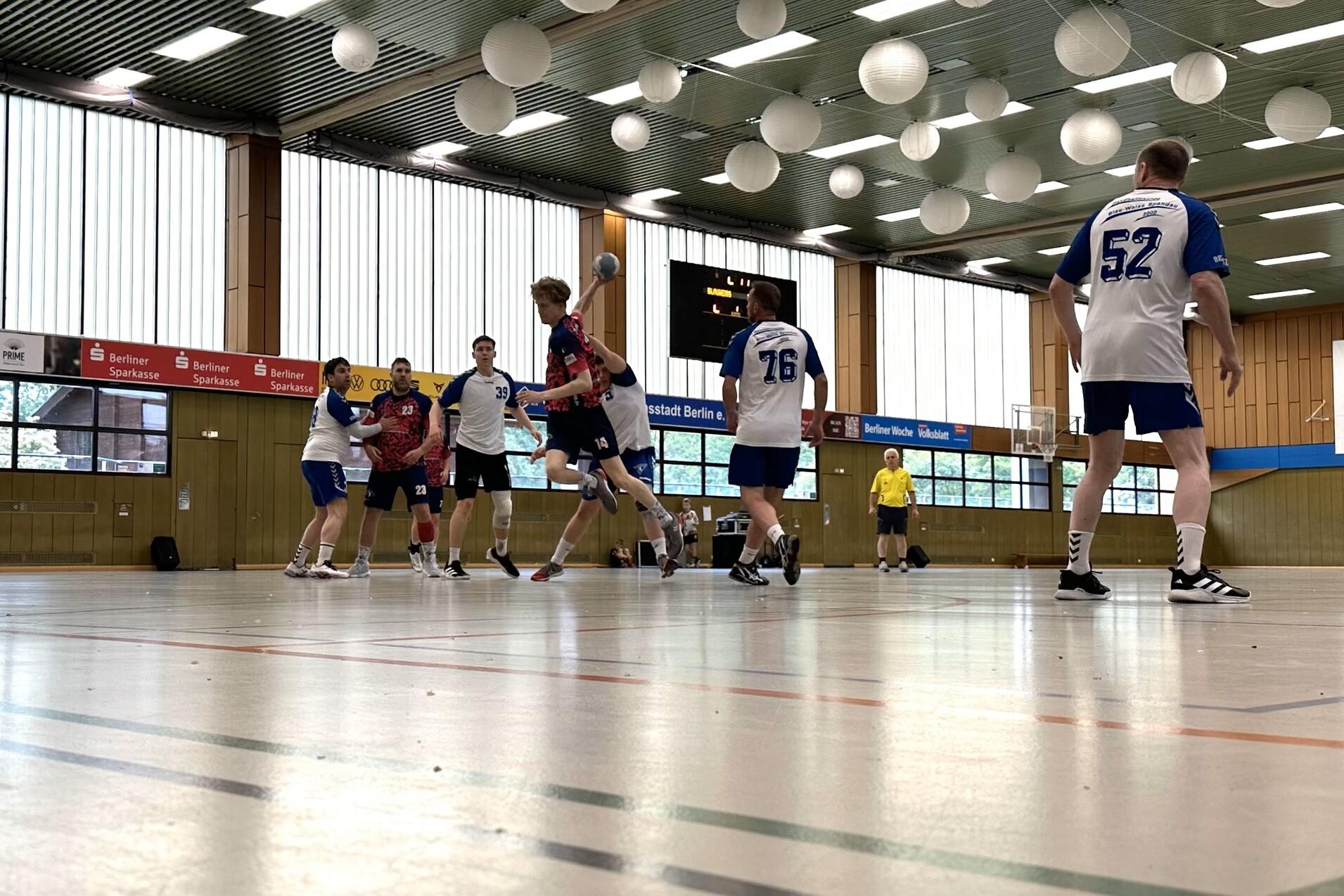 Handballspiel in einer Halle mit Spielern in wei&szlig;en und blauen Trikots, w&auml;hrend der Ball von einem Spieler geworfen wird.