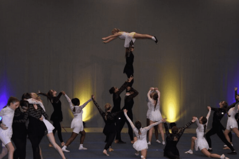 Tanzgruppe f&uuml;hrt akrobatische Elemente auf, mit einer T&auml;nzerin in der Luft &uuml;ber mehreren anderen T&auml;nzern.