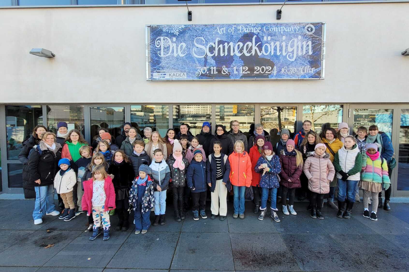 Gruppe von Kindern und Erwachsenen steht vor einem Geb&auml;ude mit Plakat f&uuml;r &bdquo;Die Schneek&ouml;nigin&ldquo;, ein Tanzprojekt.