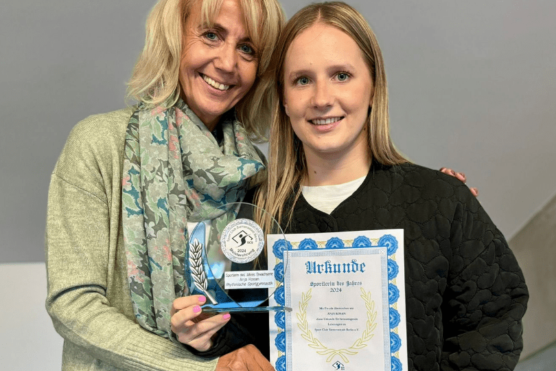 Zwei Frauen l&auml;cheln, eine h&auml;lt einen Pokal, die andere eine Urkunde, beide stehen vor einer neutralen Wand.