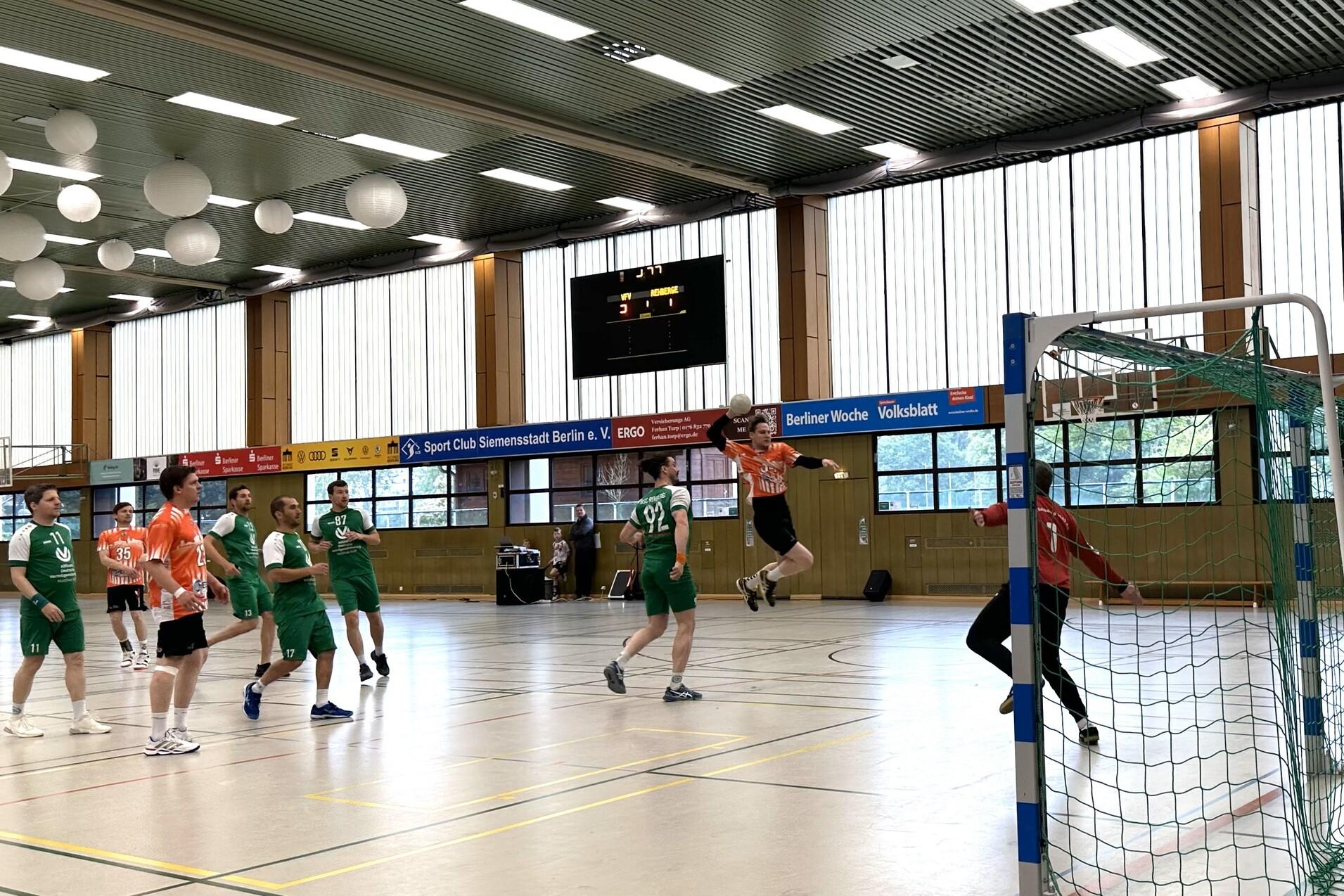 Aktion im Handballspiel: Spieler in orange springt zum Torwurf, w&auml;hrend gr&uuml;ne Spieler und Torwart beobachten.